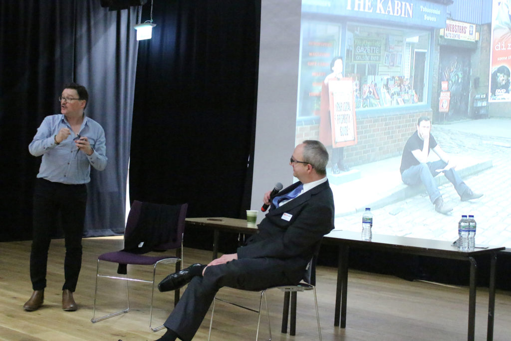 A picture showing Ian Puleston-Davies standing next to a chair and talking with a seated Ashley Fulwood from OCD UK at the 2015 York OCD UK conference