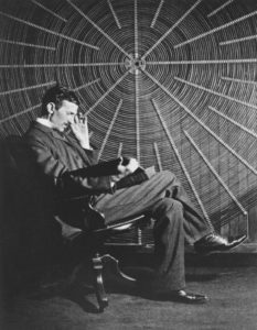 Tesla sitting in front of a spiral coil used in his wireless power experiments at his East Houston St. laboratory.