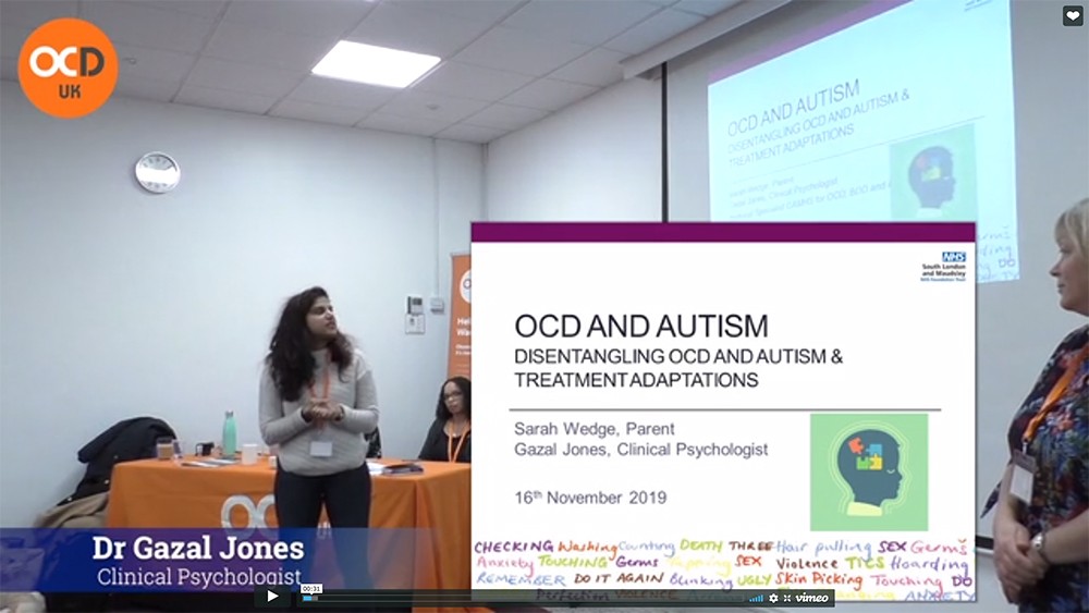 Image shows Sasha Walters talking to the audience at the 2019 OCD-UK conference looking at the projector screen alongside a screenshot of the slide.